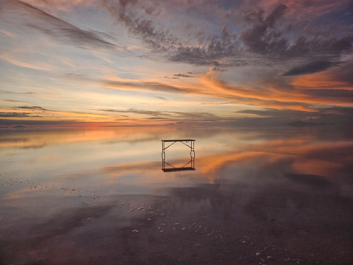 A folding table standing on a flooded salt flat at sunset, mirrored in the water.