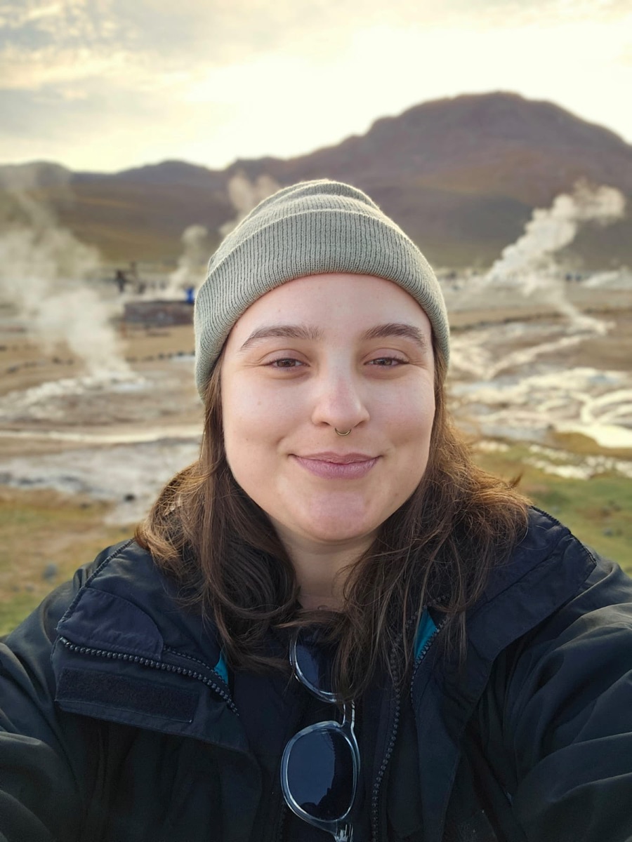 Sara smiling, wearing a beanie, with steaming geysers and mountains behind her.